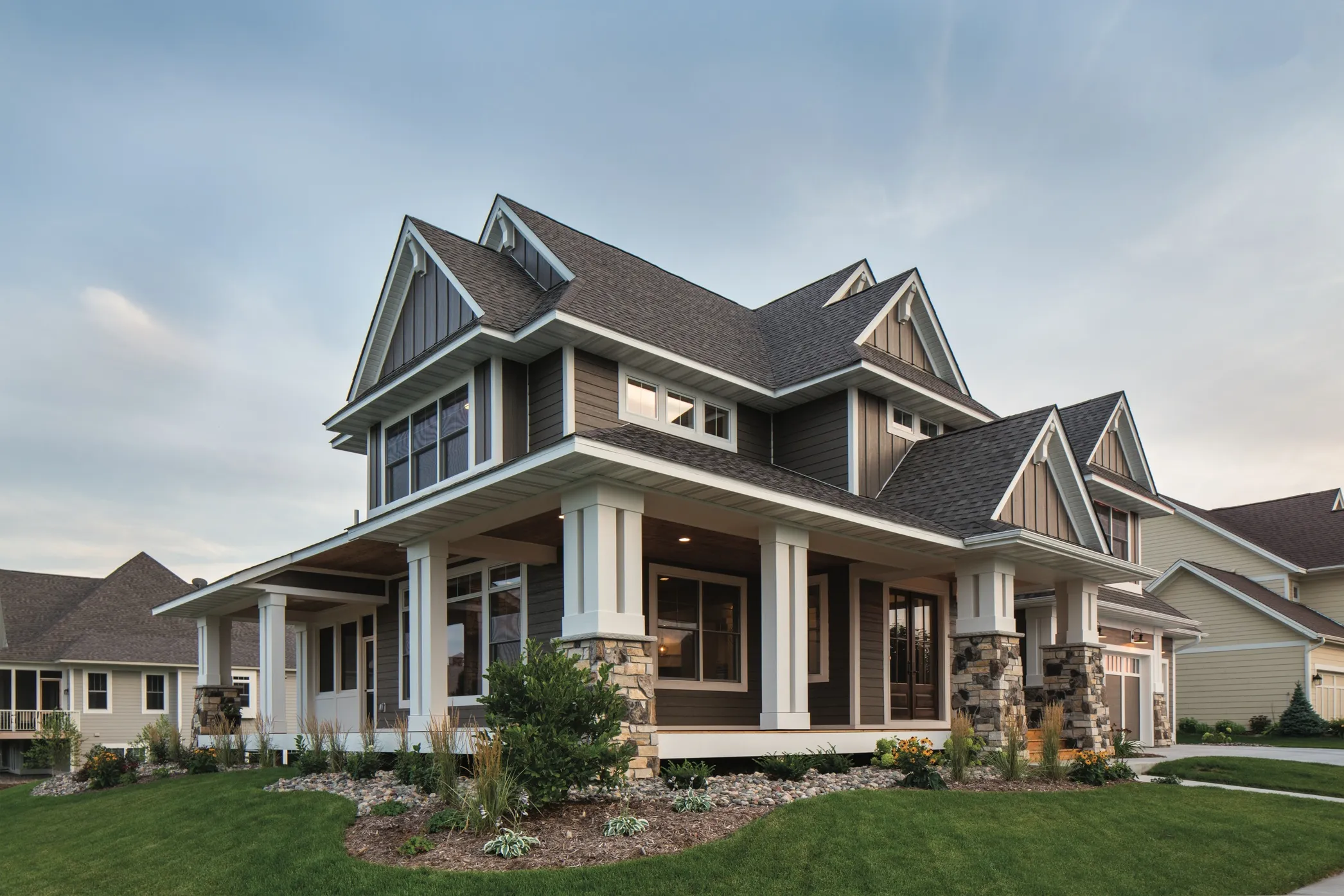 Modern two-story house with gray siding, stone pillars, wraparound porch, and manicured front lawn.